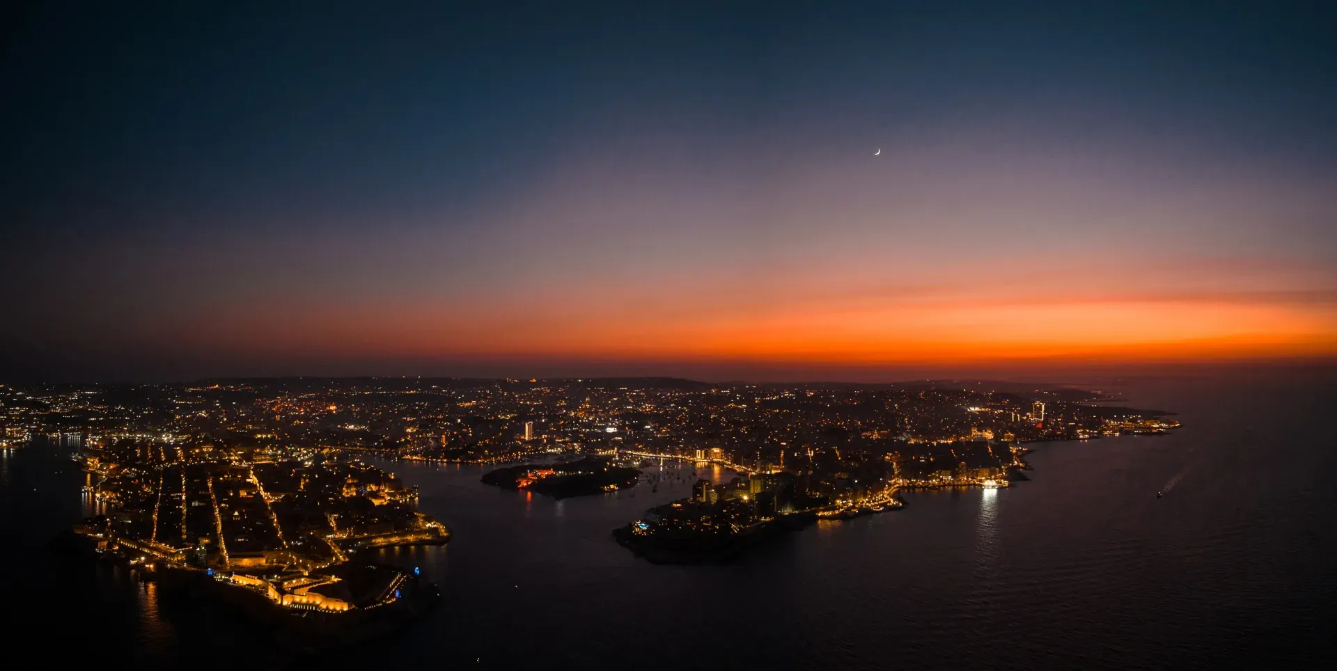 The Mediterranean coastline at dusk, harbour lights glowing beneath a deep navy and amber sky